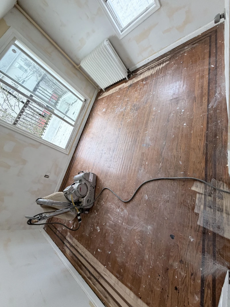 Worn floor before sanding, drum sander resting on the boards and a patch of replaced boards showing lighter along the border